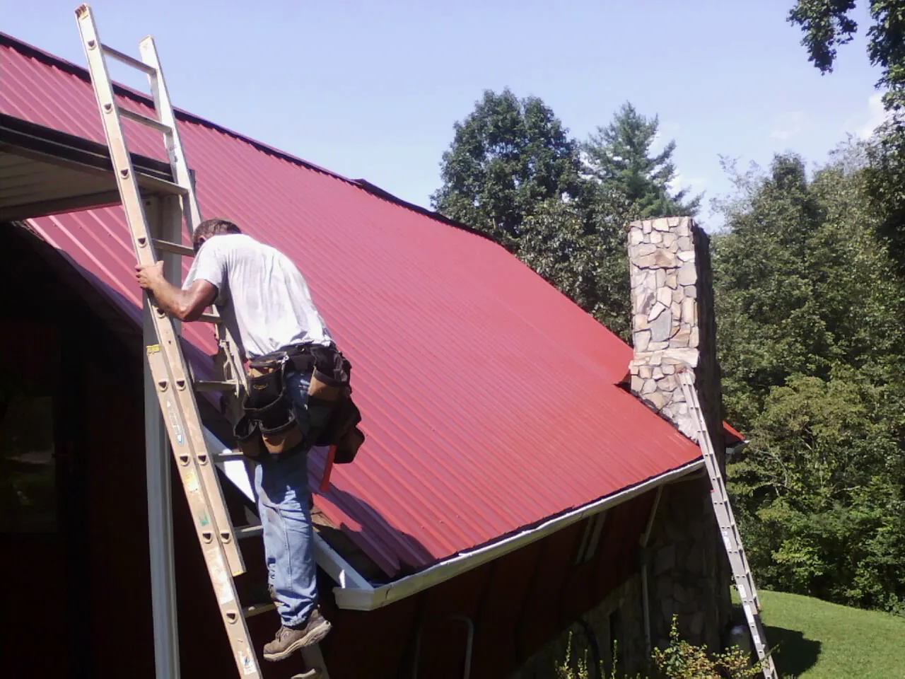 Roofer on a ladder beside a red metal roof and stone chimney — Soffit & Fascia Repair by Roofing Brothers in Center Sandwich